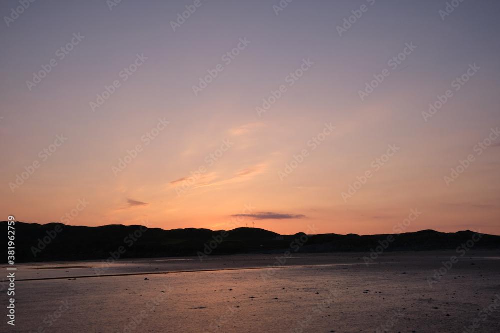 Sunset on Uisken beach in the Ross of Mull on the Isle of Mull, Scotland. Reflections of the colourful sunset are seen in the wet sand in the foreground and wind turbines are centre frame.
