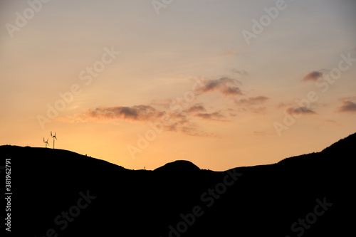 A fiery sky is lit up by the late sunset tones on a summers evening on the Isle of Mull, Scotland. Silhouetted mountains are visible and two wind turbines sit on the tops.
