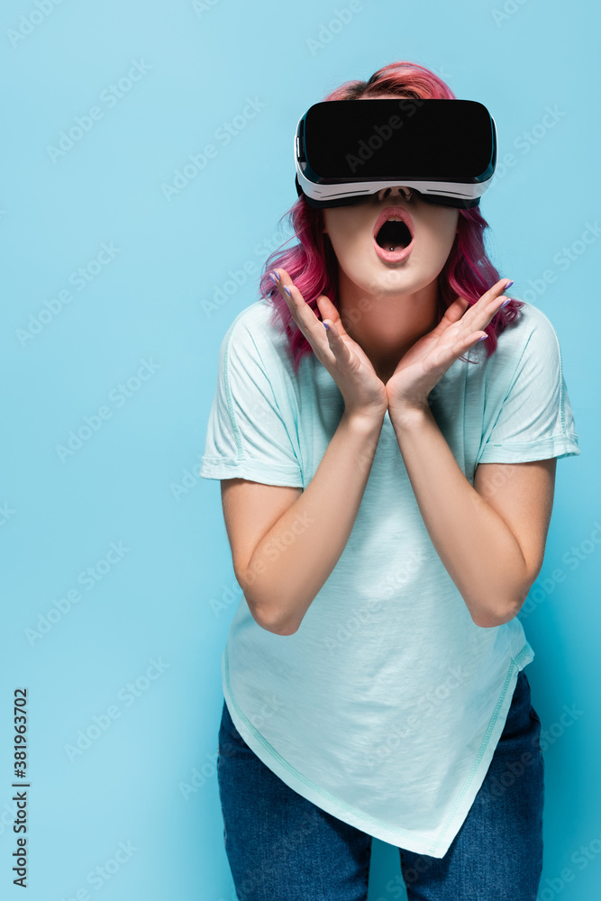 shocked young woman with pink hair in vr headset holding hands near face on blue background shocked young woman with pink hair in vr headset holding hands near face on blue background