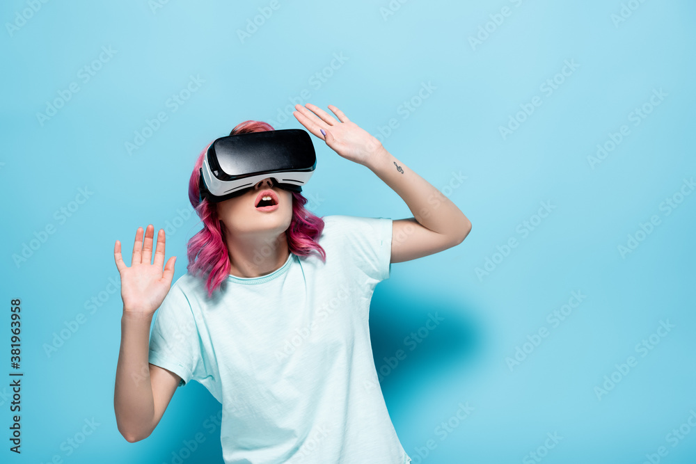 scared young woman with pink hair in vr headset gesturing on blue background scared young woman with pink hair in vr headset gesturing on blue background