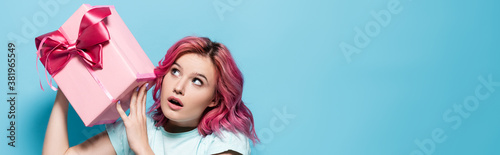 young woman with pink hair and open mouth holding gift box with bow on blue background, panoramic shot