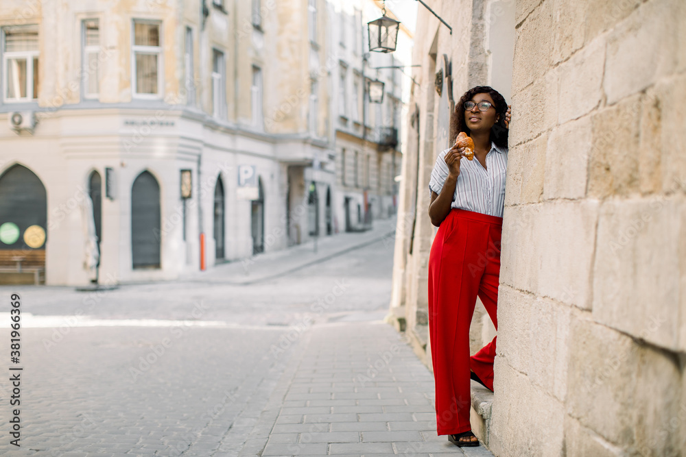 Charming young African woman in red pants, posing to camera with fresh tasty croissant, standing outdoors on the background of old vintage city buildings.