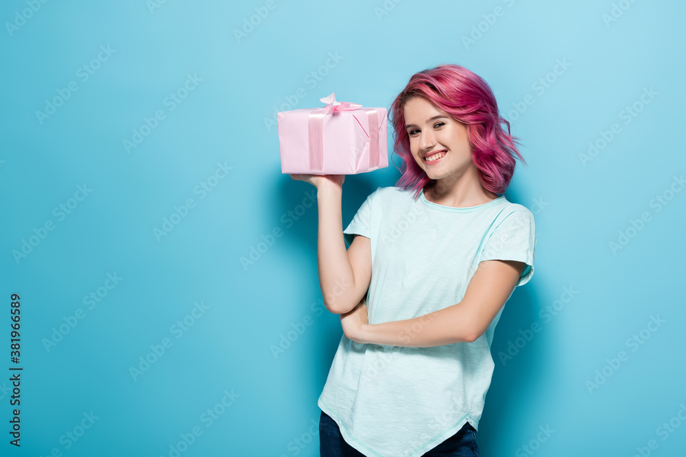 young woman with pink hair holding gift box with bow and smiling on blue background young woman with pink hair holding gift box with bow and smiling on blue background