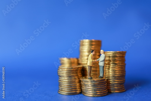 Business, Money Investment and Planning Concept. Close up of two businessman miniature people figure make hand shake on top of stack of gold coins on blue background with copy sapce.