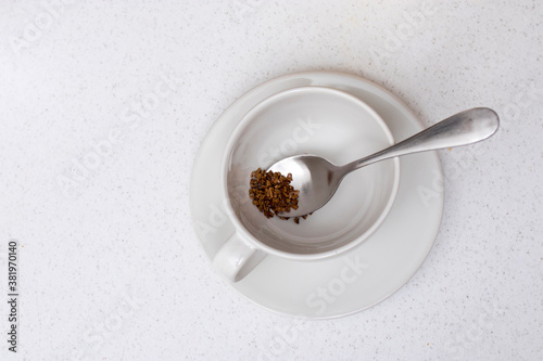Concept. Empty white ceramic cup and saucer with dry instant coffee on a spoon inside on a beige wooden table