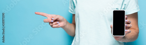 cropped view of young woman holding smartphone with blank screen and pointing aside on blue background, panoramic shot