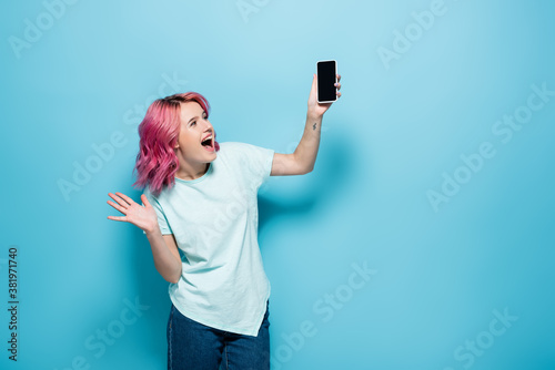 excited young woman with pink hair holding smartphone with blank screen on blue background
