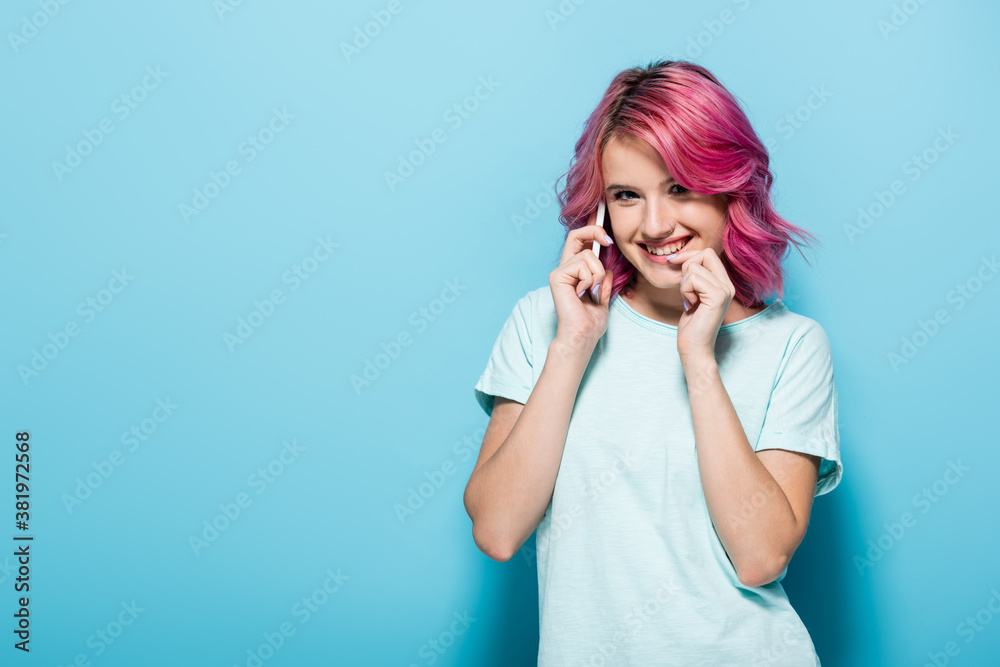 young woman with pink hair talking on smartphone and smiling on blue background