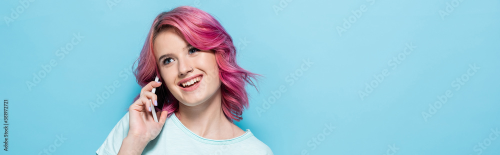 young woman with pink hair talking on smartphone and smiling on blue background, panoramic shot young woman with pink hair talking on smartphone and smiling on blue background, panoramic shot
