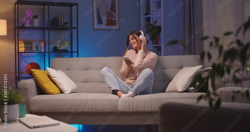 Joyful beautiful Caucasian young woman tapping on smartphone and listening to music in headphones while resting on sofa at home. Happy female enjoying songs from cellphone in room. Leisure concept Joyful beautiful Caucasian young woman tapping on smartphone and listening to music in headphones while resting on sofa at home. Happy female enjoying songs from cellphone in room. Leisure concept