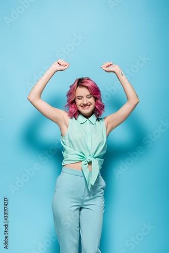 young woman with pink hair and hands in air smiling on blue background © LIGHTFIELD STUDIOS