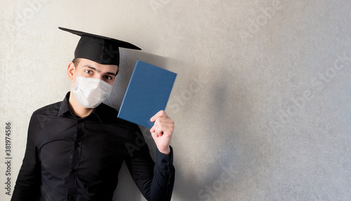 Portrait of a young student in a graduation cap and protective mask with a diploma in his hand, on a gray background, banner. The concept of distance education in the context of the coronavirus pandem