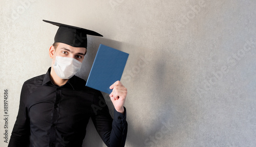Portrait of a young student in a graduation cap and protective mask with a diploma in his hand, on a gray background, banner. The concept of distance education in the context of the coronavirus pandem photo