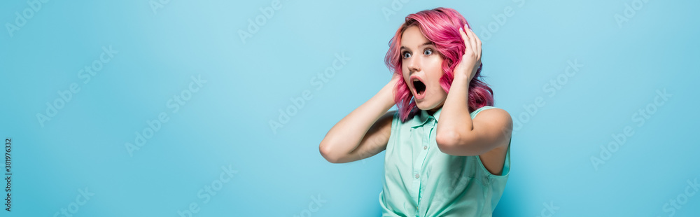 shocked young woman with pink hair and open mouth holding head on blue background, panoramic shot