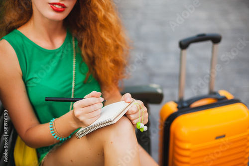 young beautiful sexy woman, hipster outfit, red hair, traveler, green top, orange suitcase, making notes, travel diary book