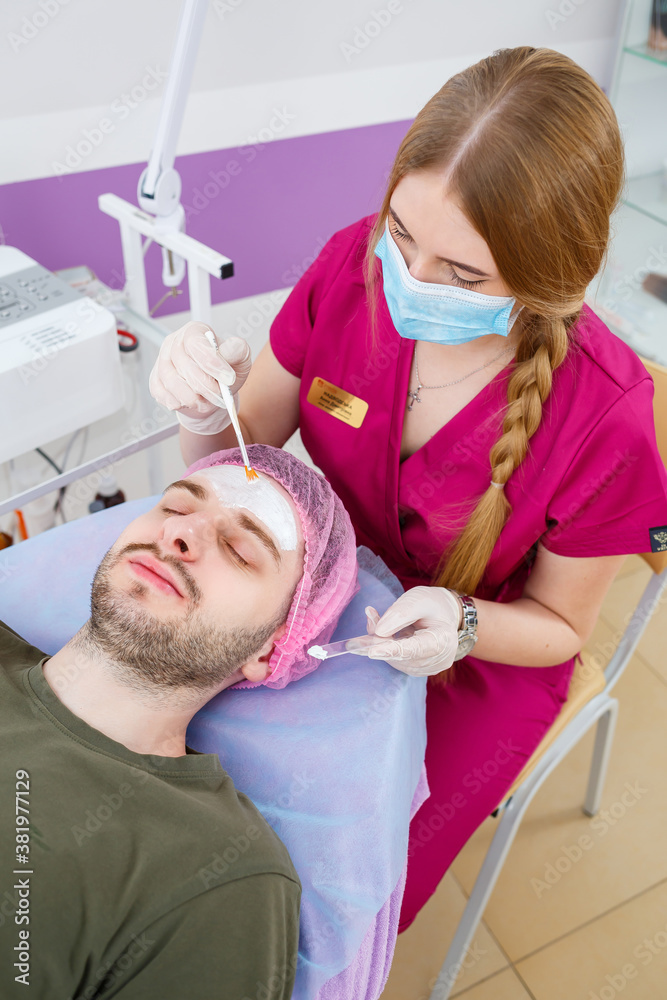 Cosmetologist is applying white moisturizing mask on man's face using brush in beauty clinic, face closeup. Beauty industry concept. Guy is lying on couch. Beautician woman making spa procedure. Cosmetologist is applying white moisturizing mask on man's face using brush in beauty clinic, face closeup. Beauty industry concept. Guy is lying on couch. Beautician woman making spa procedure.