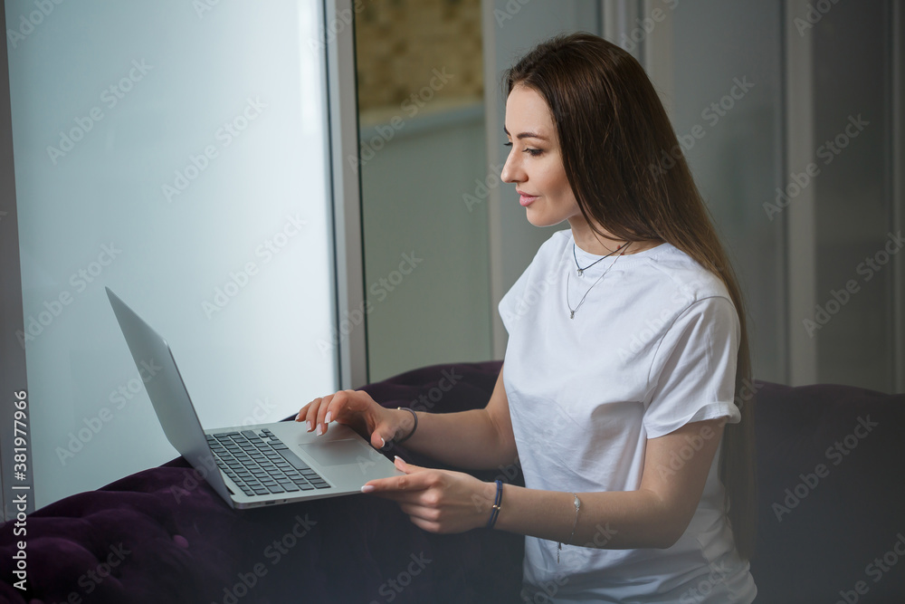 Naklejka premium A young woman is sitting on the couch and working on a laptop remotely from work. Girl with a computer on her knees looking at the monitor