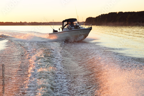 Fast gliding motor boat in water foam track on beautiful sunset background, bright orange sunshine in calm water on forest on Horizon at Sunny summer evening bow side front view from powerboat transom
