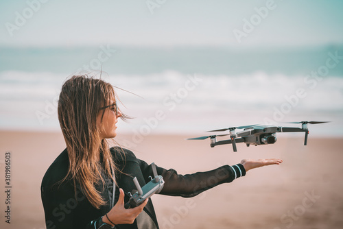 Fotografi Young modern lady pilot in black jacket lands drone on her hand with remote controller near the ocean