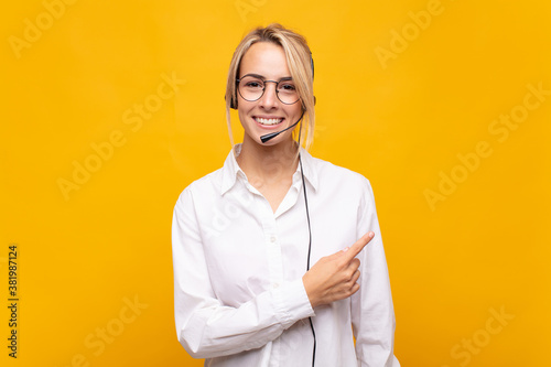 young woman telemarketer smiling cheerfully, feeling happy and pointing to the side and upwards, showing object in copy space