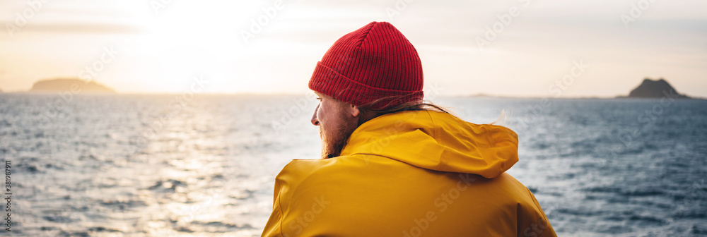 Wide image. Alone man traveling on ship and looking at sunset sea and foggy mountain on skyline. Hipster traveler wearing yellow raincoat and red hat enjoying beautiful ocean after storm - obrazy, fototapety, plakaty 
