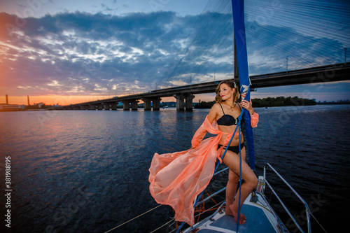 Young beautiful girl standing on the yacht in pink dress. Yacht is sailing toward to the sunset. against the background of the bridge