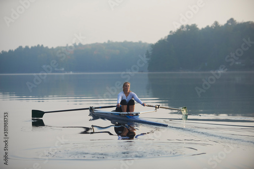Middle aged woman rowing on calm autumn lake at early morning with mist over water.