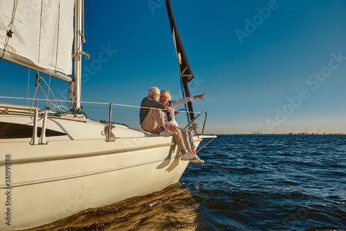 Sailing. Happy retired family couple sitting on the side of a sail boat or yacht deck floating in a calm blue sea, pointing at the horizon and enjoying amazing sunset