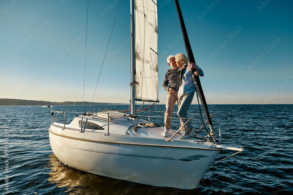 Full length of a happy senior couple in love, man and woman standing on the side of yacht deck floating in sea, they are hugging and kissing while having boat trip Full length of a happy senior couple in love, man and woman standing on the side of yacht deck floating in sea, they are hugging and kissing while having boat trip