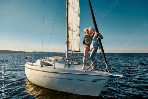 Full length of a happy senior couple in love, man and woman standing on the side of yacht deck floating in sea, they are hugging and kissing while having boat trip