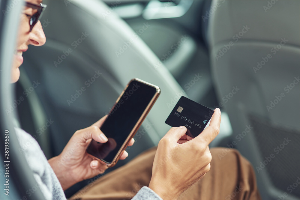 Close up shot of smiling business woman using her smartphone and credit card to buy something while sitting on back seat in the car Close up shot of smiling business woman using her smartphone and credit card to buy something while sitting on back seat in the car