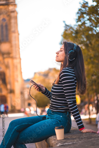 Lifestyle, Caucasian brunette enjoying listening to music in the city. Relaxing with a take away coffee jungo to a beautiful church in the park. Black sweater with white stripes
