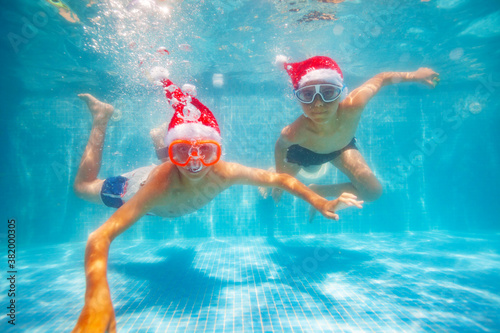 Two cool boys in scuba masks dive and swim underwater wearing Santa Claus hat in the pool