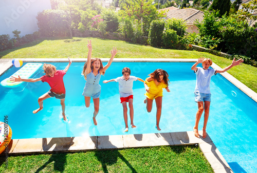 Group of happy teen children in mid air fall into pool water hands up view from above