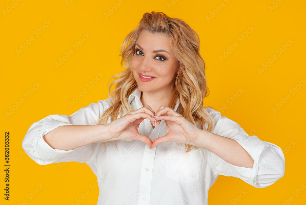 Blondine woman in white shirt, middle aged European girl, showing heart gesture, recognizing sympathy, look smiling friendly at the camera, girlfriend expresses true feelings, on yellow background Blondine woman in white shirt, middle aged European girl, showing heart gesture, recognizing sympathy, look smiling friendly at the camera, girlfriend expresses true feelings, on yellow background
