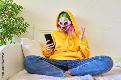 Halloween party at home, teenage girl in scary hoodie mask with smartphone