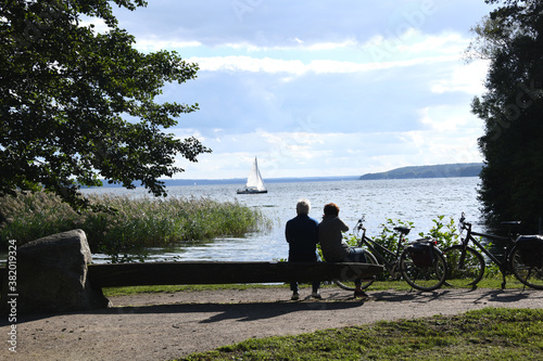 Entspannung am Tollensesee in Neubrandenburg. Ein Paar bei der Rast beobachtet ein Segelschiff am Ufer. Sie entspannen sich auf einer Bank nach einer Fahrradtour. 