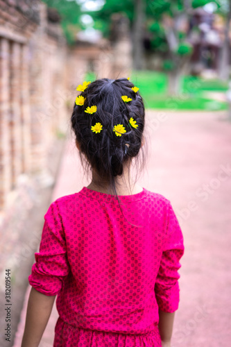 Back view of little asian girl in red dress with beautiful hair and yellow flower walking in the park