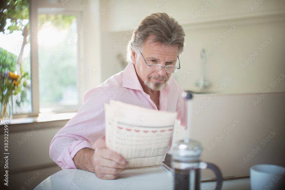Senior man reading newspaper at laptop at morning kitchen table Senior man reading newspaper at laptop at morning kitchen table