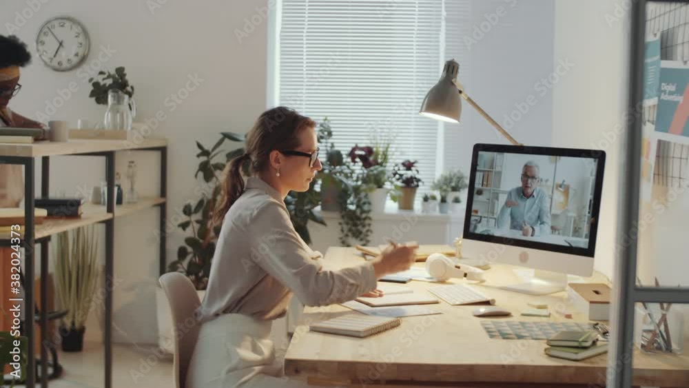 Young caucasian woman discussing business with senior male colleague via online video call on computer and taking notes in notepad while her black female coworker standing in the background