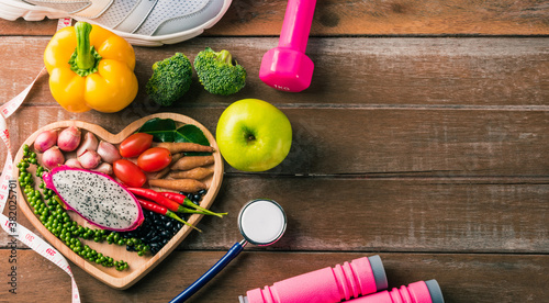Top view of fresh organic fruit and vegetable in heart plate, shoes, sports equipment and doctor stethoscope, studio shot on wooden gym table, Healthy diet vegetarian food concept, World food day