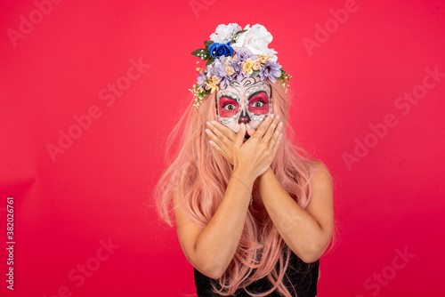 young beautiful woman wearing halloween make up over red background shocked covering mouth with hands for mistake. Secret concept.