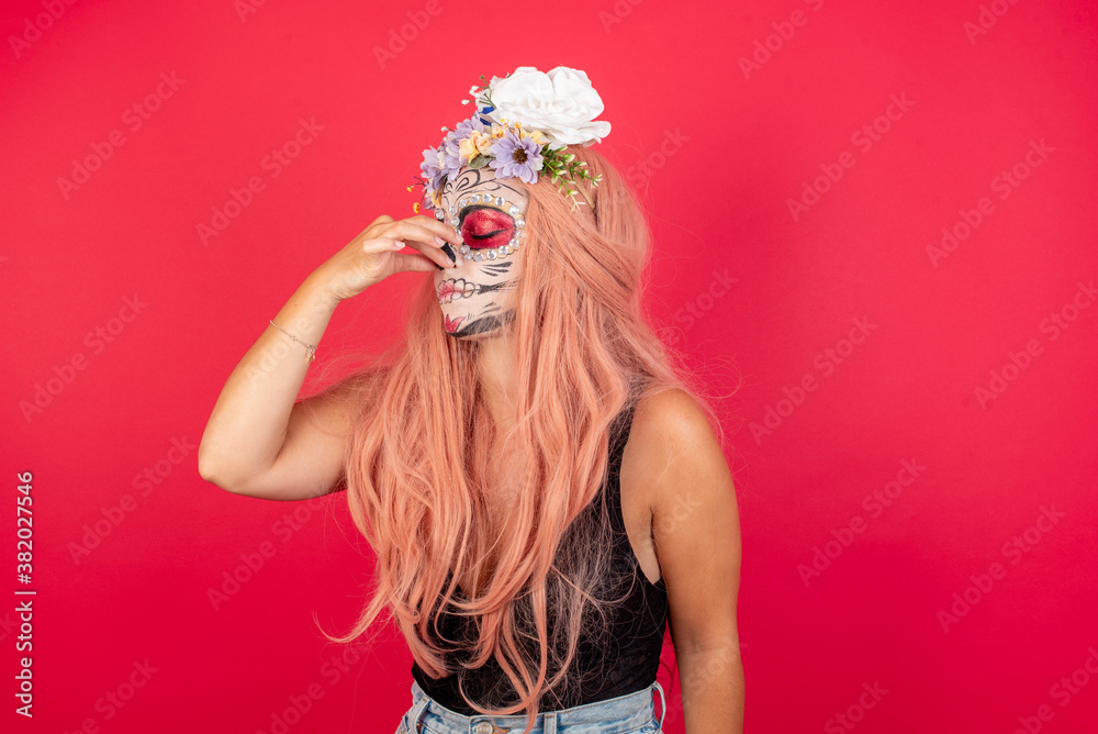 young beautiful woman wearing halloween make up over red background smelling something stinky and disgusting, intolerable smell, holding breath with fingers on nose. Bad smell