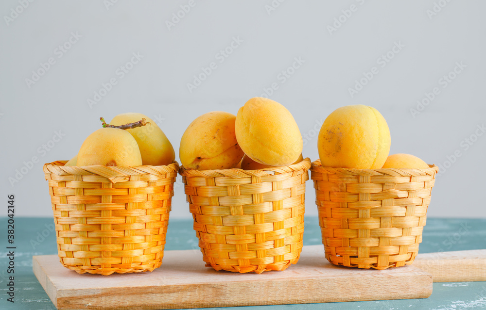 Apricots with cutting board in baskets on plaster and white background, side view.