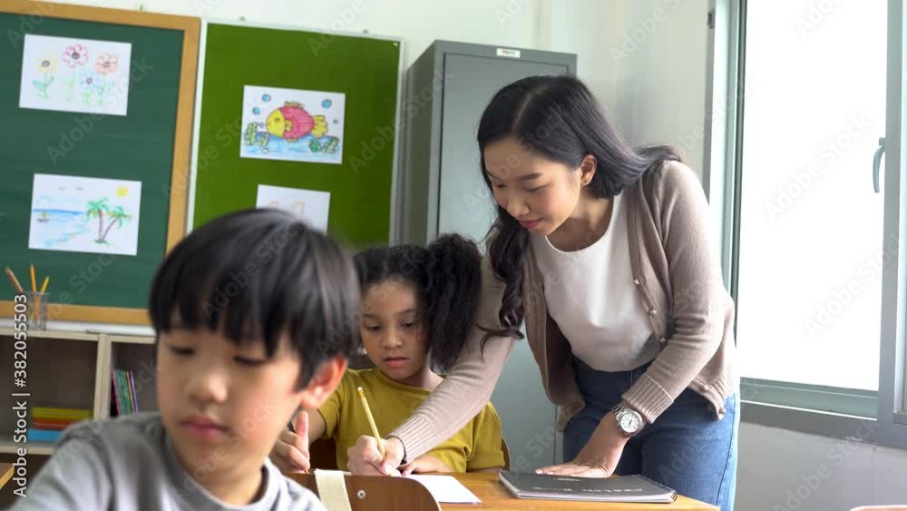 Asian school teacher assisting students in classroom. Young woman working in school helping boy with his writing, education, support, care. Asian school teacher assisting students in classroom. Young woman working in school helping boy with his writing, education, support, care.