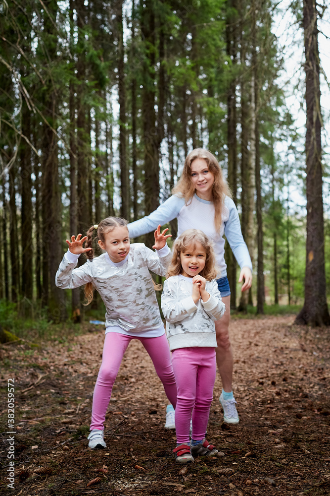 Mother and her child sister girls playing and having fun together on walk in forest outdoors. Happy loving family posing on nature landscape with with pine trees. Mother and her child sister girls playing and having fun together on walk in forest outdoors. Happy loving family posing on nature landscape with with pine trees.