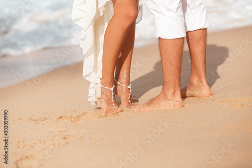 Brides feet with beautiful jewelry ad groom legs on sandy beach, just married couple walk by the sea. Wedding near ocean
