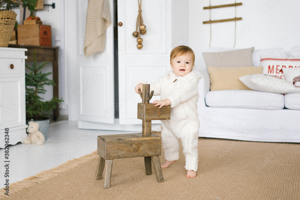 A one-year-old child in a soft jumpsuit stands next to a wooden deer toy in the living room, decorated for Christmas or New Year A one-year-old child in a soft jumpsuit stands next to a wooden deer toy in the living room, decorated for Christmas or New Year