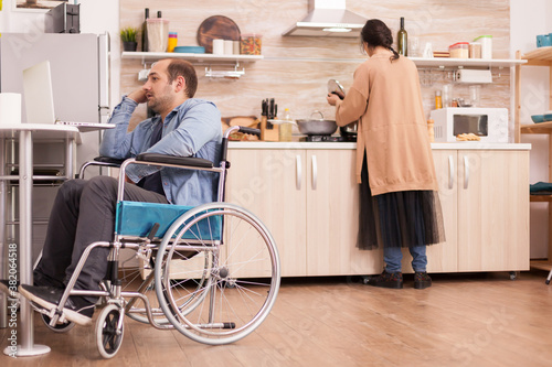 Handicapped man in wheelchair using laptop in kitchen and wife is preparing meal. Disabled paralyzed handicapped man with walking disability integrating after an accident.