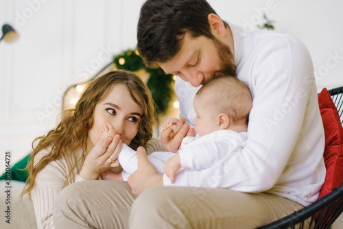 Mom and dad kiss their baby in the living room, decorated for New Year and Christmas in the bedroom or living room. Family emotional portrait